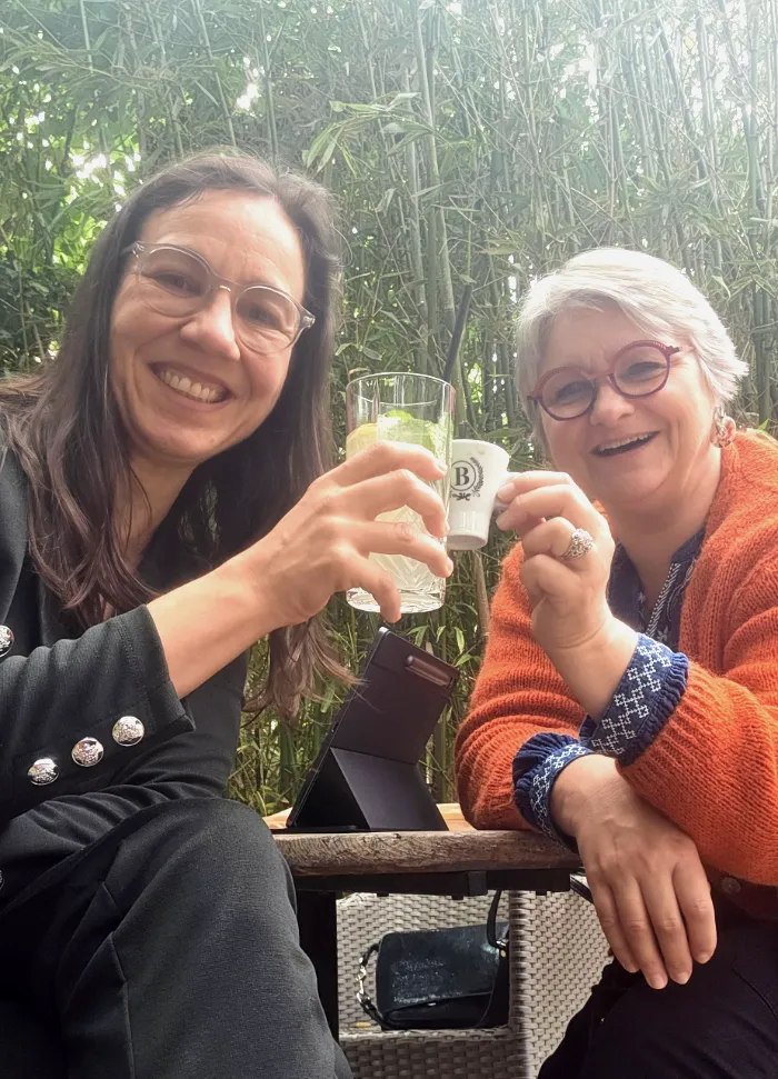 Rita Cuzzupi et Sylvie Boczkowski trinquent joyeusement avec un verre de ginger beer et un café, sur une terrasse arborée